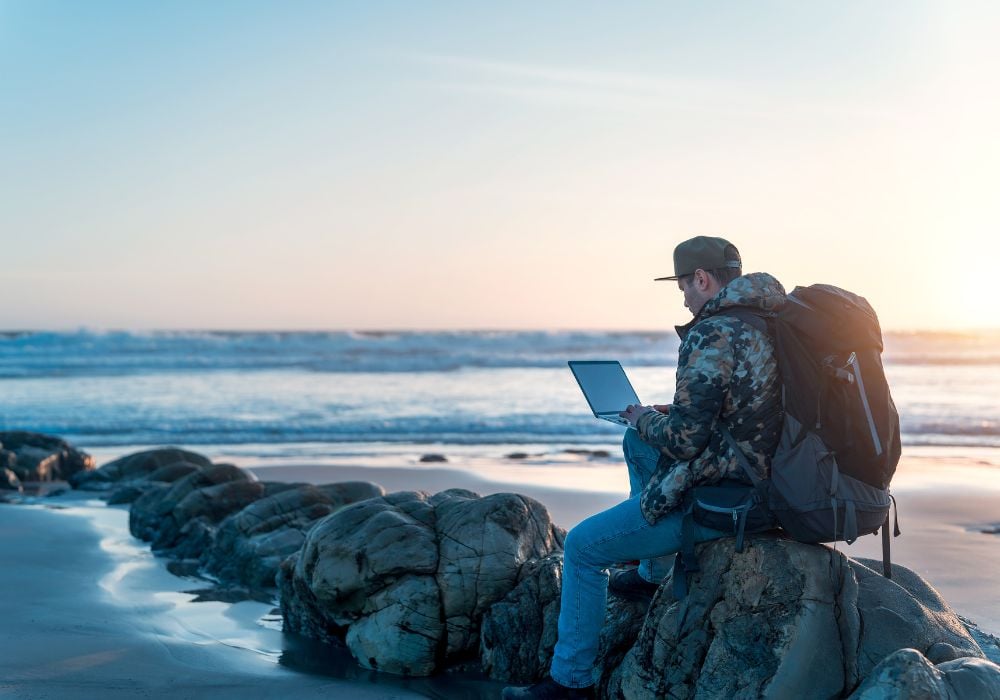 The digital nomad is sitting on a rock on the beach, working with his laptop on his backpack.