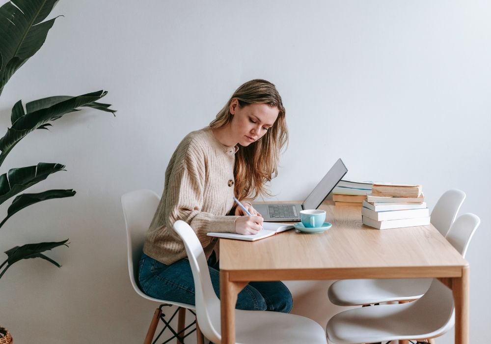 A woman is sitting on a wooden chair and writing a standout house-sitting profile on her laptop.