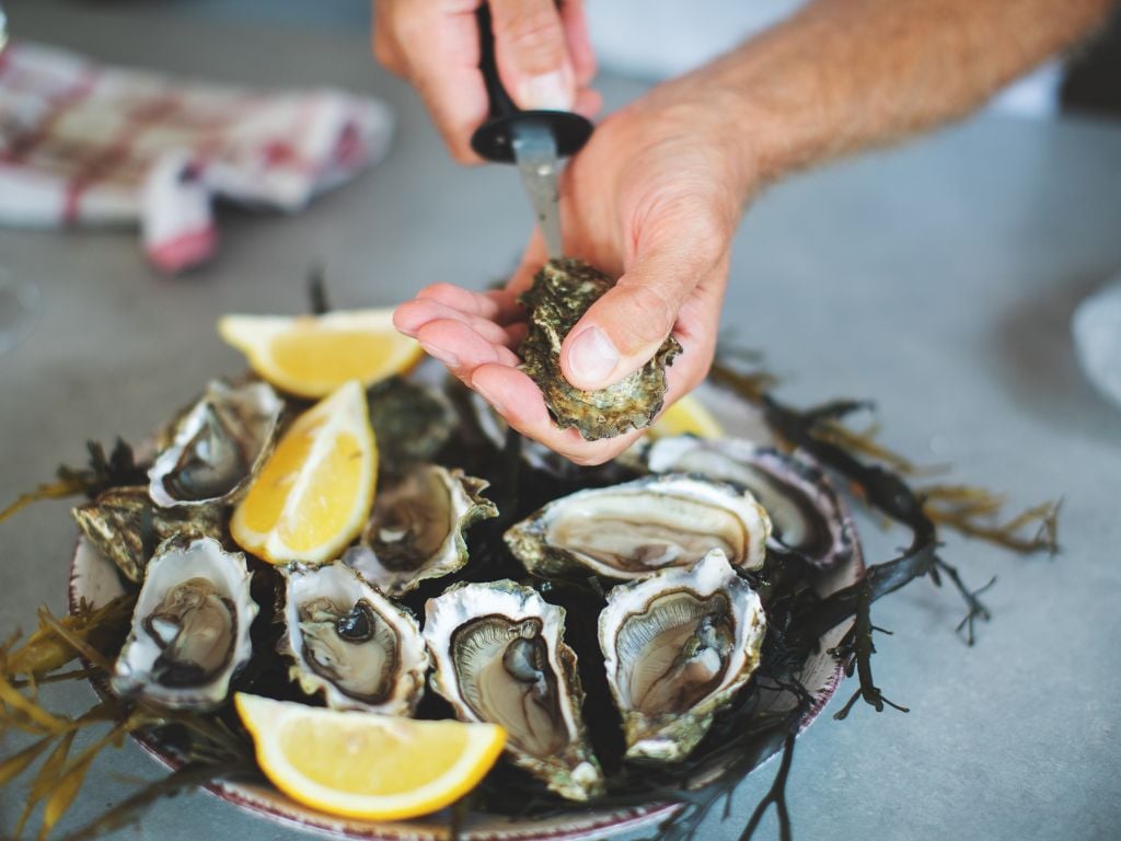 man shucking an oyster with a plate of oysters and lemons