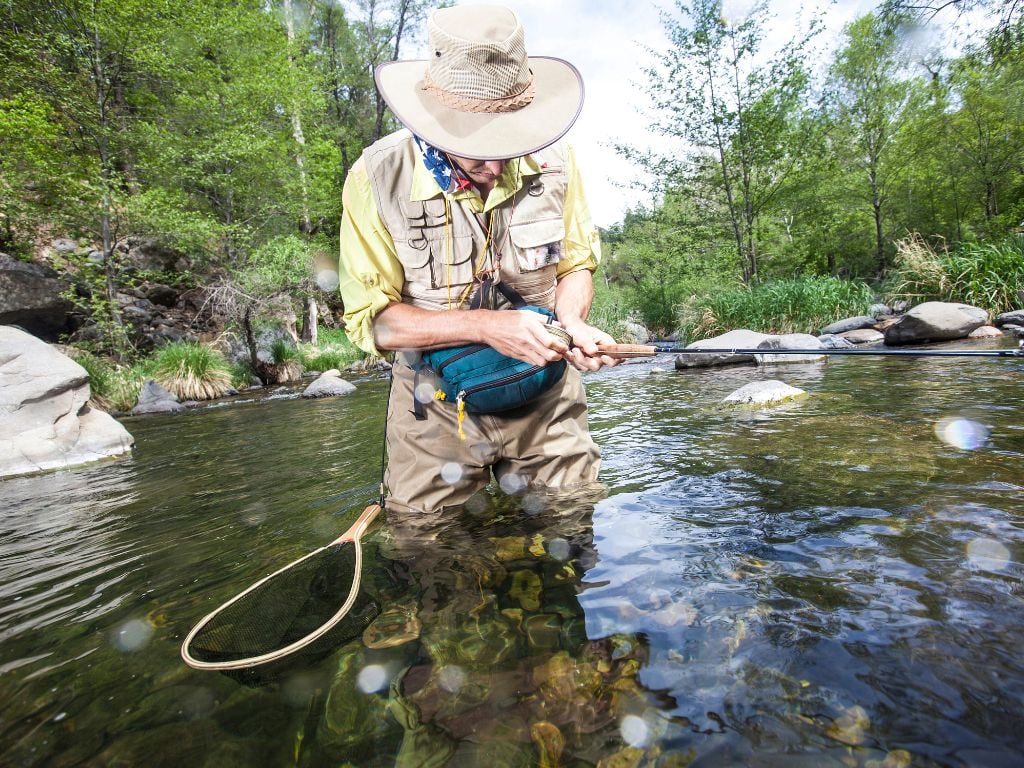man standing in a river with a fly fishing rod surrounded by trees