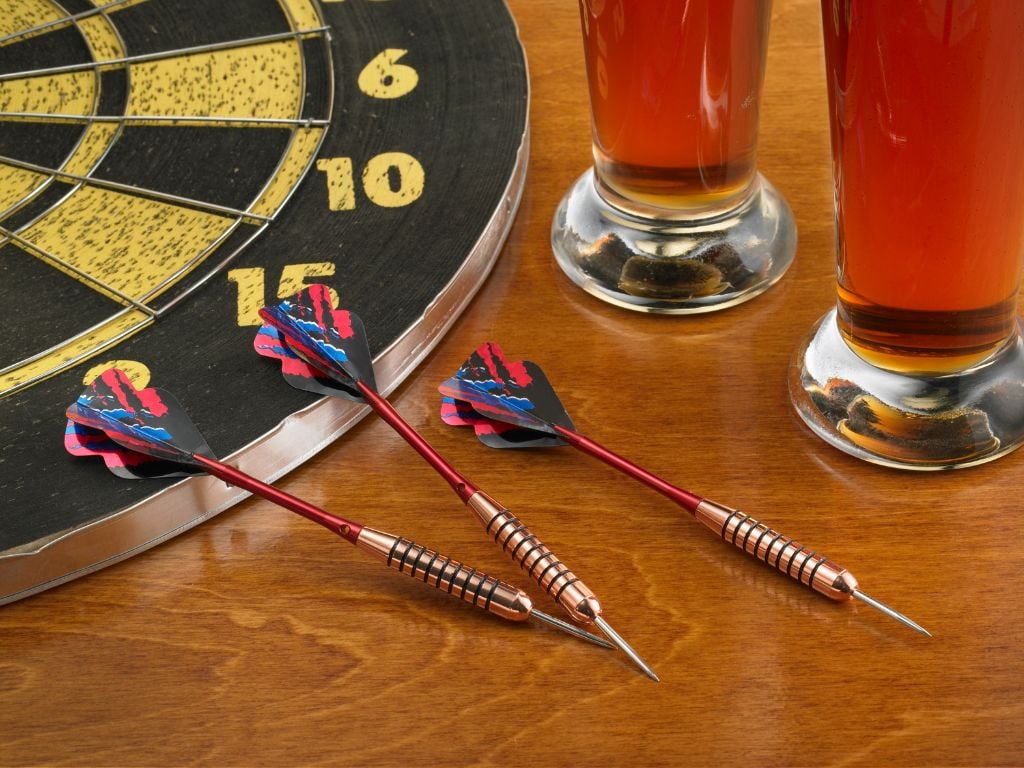 3 darts next to a dart board with 2 beers