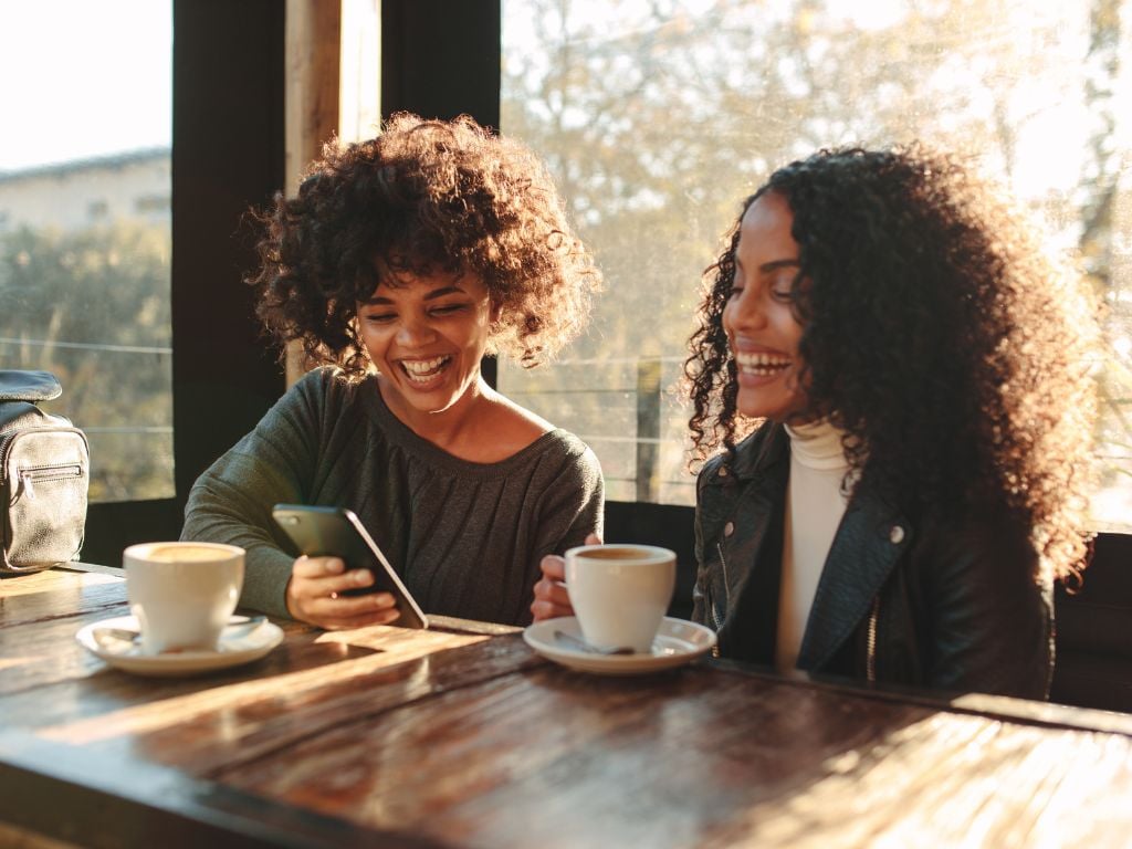 two women sitting at a table looking at an iphone with coffee cups