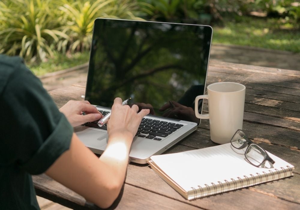 woman writing on a laptop outside at a desk with a notepad, her glasses and a coffee mug