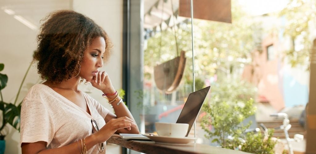 A woman using laptop and a cup of coffee on the side with plants around the cafe.