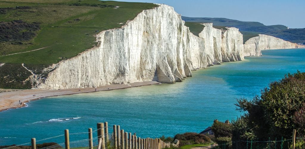 Seven Sisters and Friston Forest, a white high cliff and the green grasses above it. Beneath the cliff is an ocean 