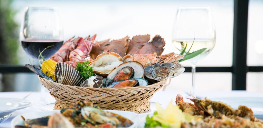 A variety of seafood in a basket served with two glasses of wine.