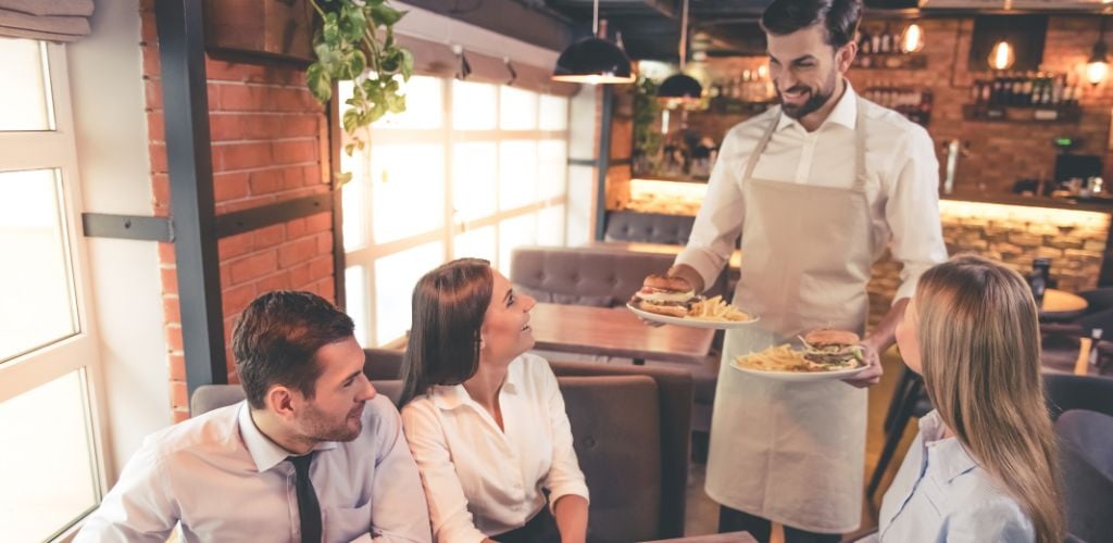 Two women and a man are delighted to receive their order, and a waiter stands ready to serve their food.