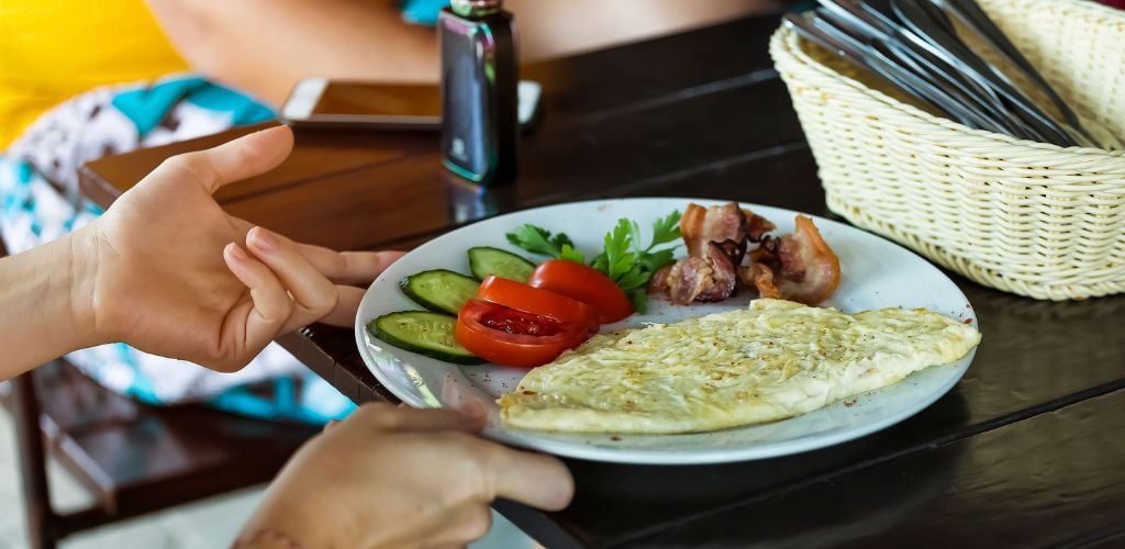 A plate containing omelet, bacon, tomato, cucumber and parsley. Two hands touching the plate which is on the table with a mobile phone in the background.