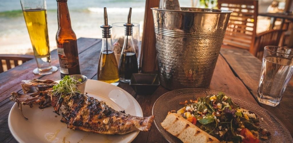 A plate of grilled fish and a plate of salad, a bucket of beer and some condiments and a glass of water on the table with sea beach in the background.