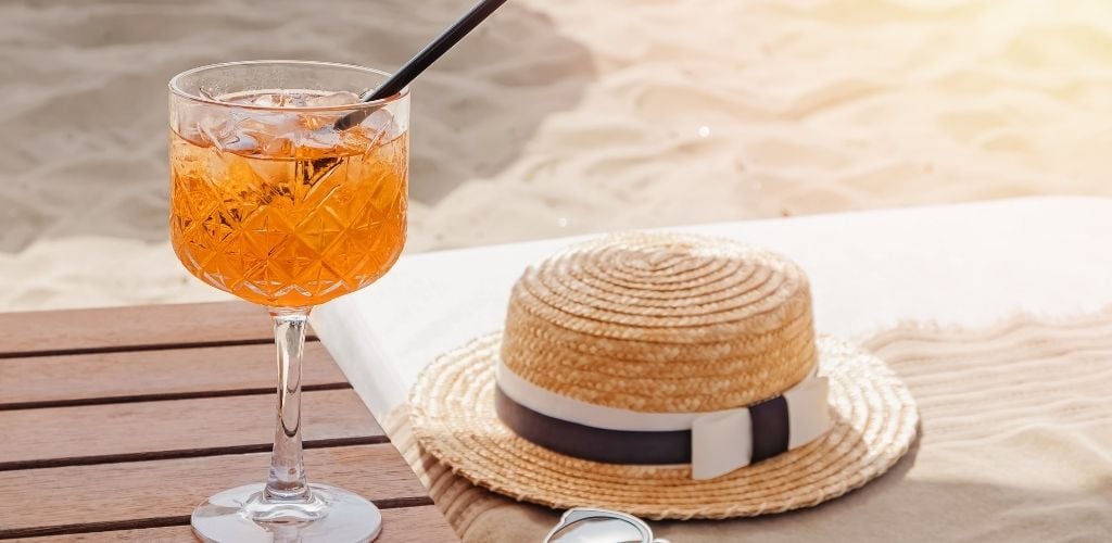 A glass of alcoholic drink and a summer cup. with sand in the background. 