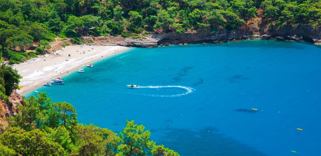 Blue beach with white sand surrounded by green mountains and there are small boats floating. 