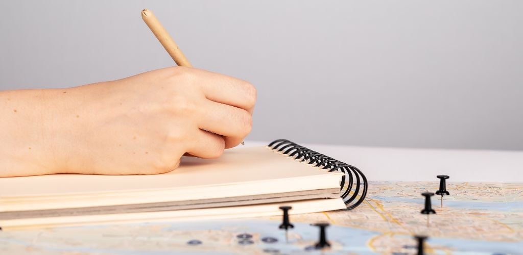 There is a hand holding a pen and writing something in a notebook, as well as a map with black pins on it. 
