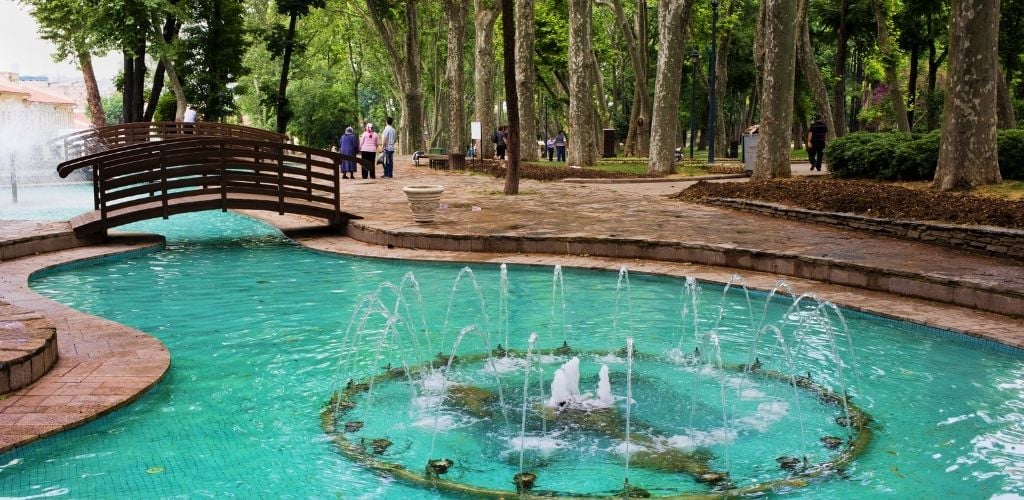 A small fountain with a mini bridge in the background and a couple of people around. 