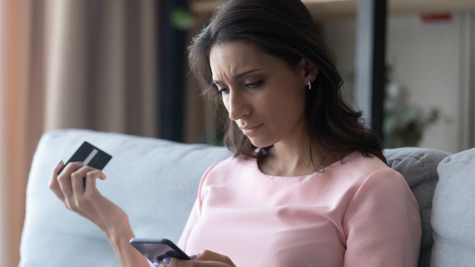 A concerned woman checks her phone while holding a debit card