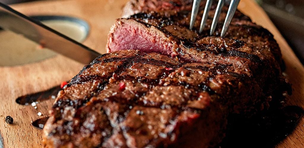 On a wooden plate, a steak is sliced with a fork and knife. 