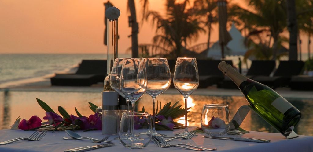 A set of six wine glasses, a bottle of wine, and cutlery on a white table at sunset, with a pool and beach in the backdrop. 