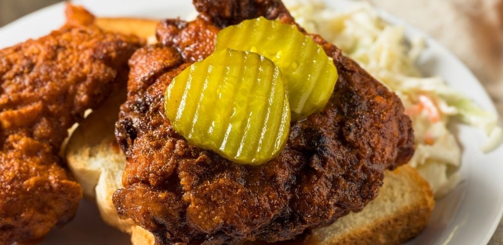 a bread and top of Nashville hot chicken and top of two pieces of pickles with a side dish of coleslaw.