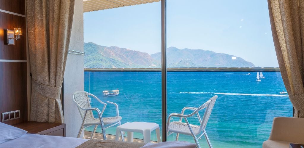 Capture the interior of a hotel room with a bed, two chairs, and a little table on the veranda with a view of a gorgeous beach, mountain, and three small boats floating in the middle of the sea outside the window.