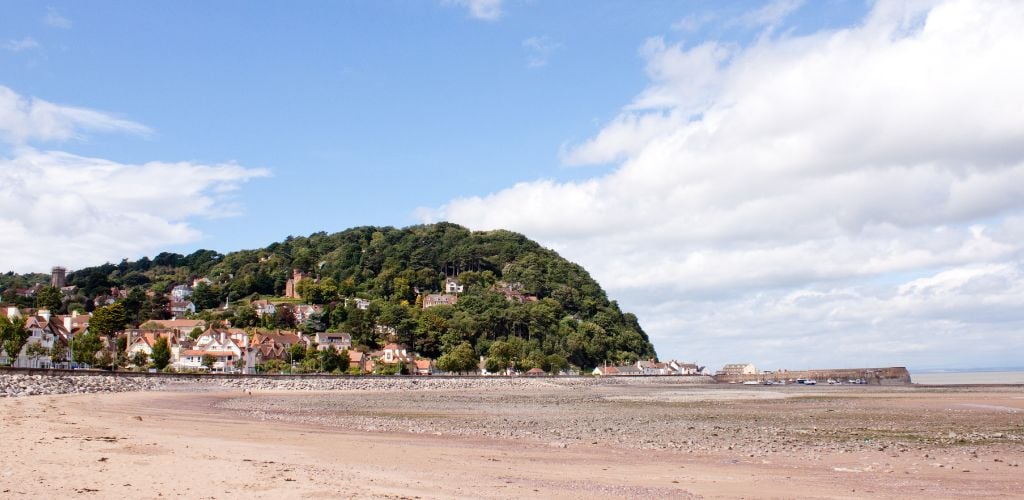 A large green mountain with residences on top. A low tide beach scene with a cloudy sky. 