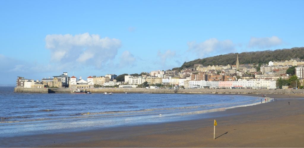 A beach scene with wavy sea, surrounded by buildings and mountains. A tiny stick with a yellow flag stands at the coast, and there are a few people walking along it. 