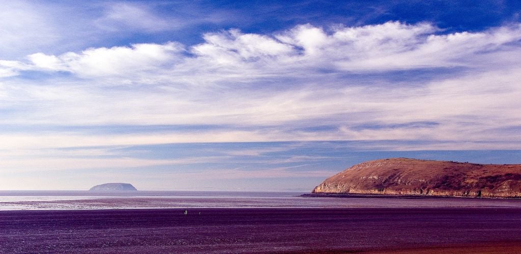 A beach view with a dry brown mountain in the background, calm sea water, and an island in the middle of the sea. The sky is cloudy. 