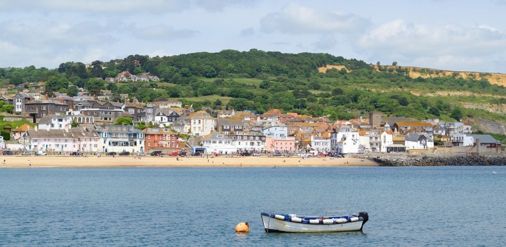 A beach view with a small boat in the center of the water, with building structures and a green mountain in the distance. 