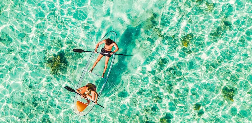 Young couple paddling on glass bottom kayak in tropical ocean. 