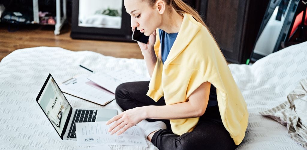 A woman is sitting on the bed with a laptop and resume. On the screen of the laptop, there is the text "Job Search"