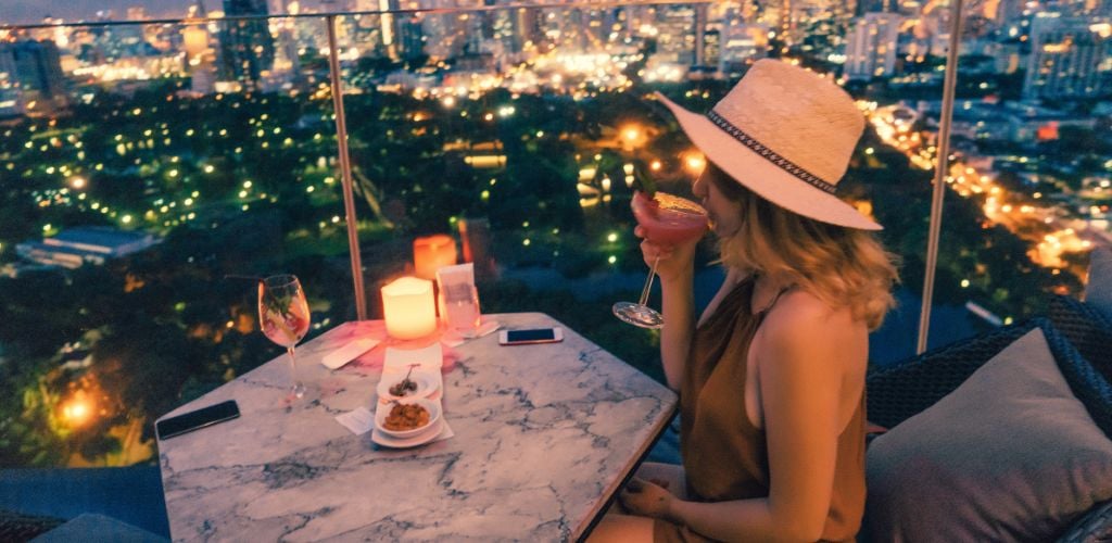 A woman wearing a summer hat and having a cocktail on a rooftop bar with a view of the city lights. 