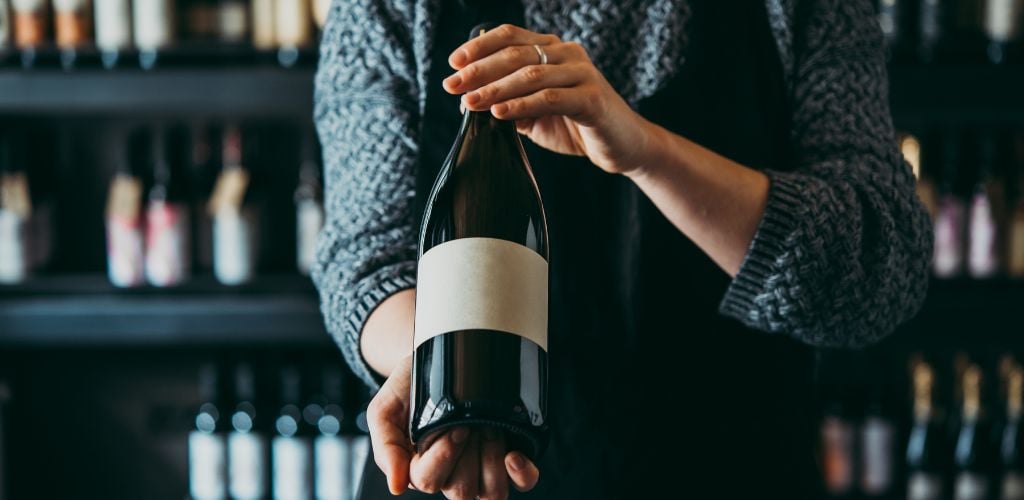 A man carrying a bottle of wine in front of a wine cabinet. 