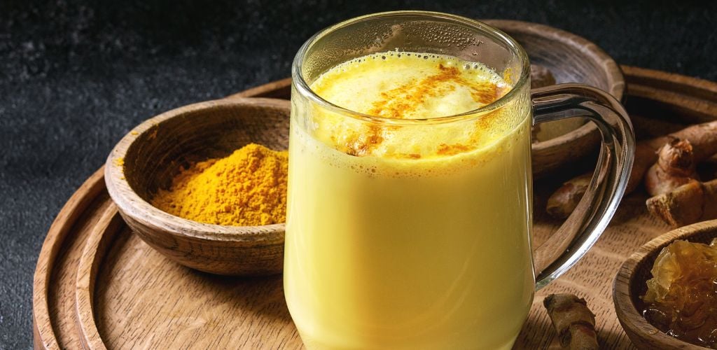 Cup of ayurvedic drink golden milk turmeric latte with curcuma powder on a round wooden tray and ingredients above over black texture background