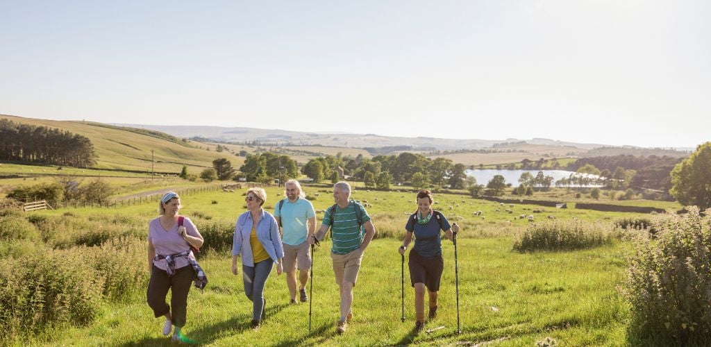 A mature group of friends are out walking in the countryside with a stunning view behind them. 