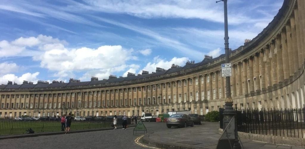 It is a long and curved historic building with a few people walking and cars part at the front.