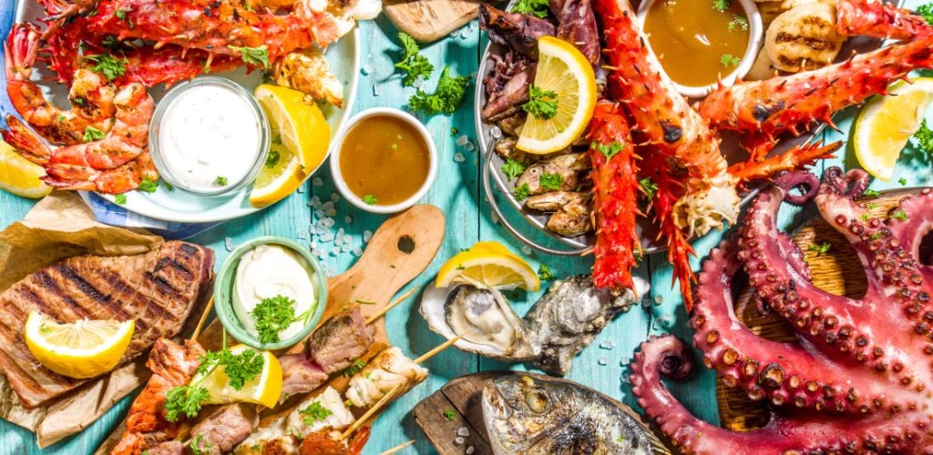 Assortment of various barbecue Mediterranean grill food- fish, octopus, shrimp, crab, seafood, mussels with kebab, sauces, light blue sunny wooden background.