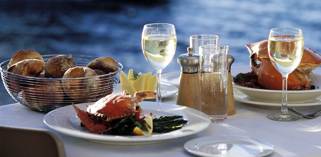 A waterfront dining, there a two plates of crab, two glasses of wine, a bowl of bread a small plate of cheese, and two bottles of condiments on the table. 