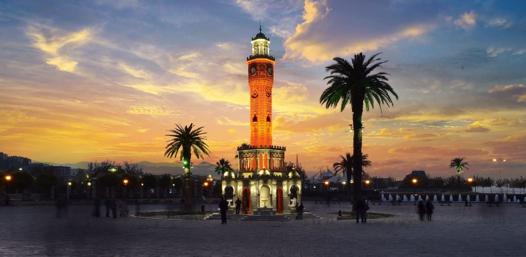 A tower of Izmir clock stands at the center of the park with trees and some people walking around. 