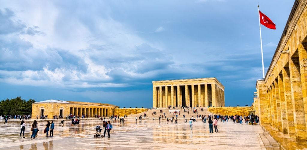 An open walking ground with crowded people, a historical of Ataturk Museum, a flag of Turkey and cloudy blue sky. 