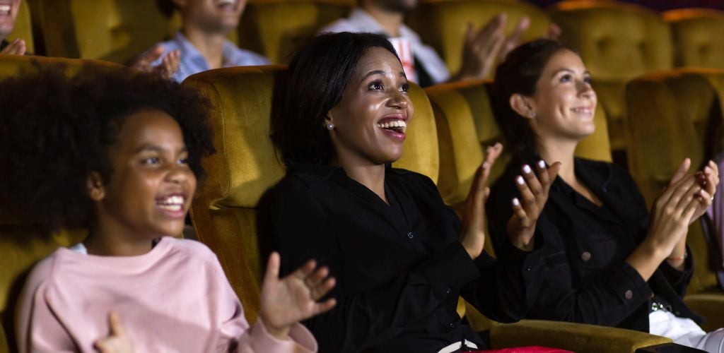 Excited black women and girls audience smiling and clapping hands while sitting amidst people. 
