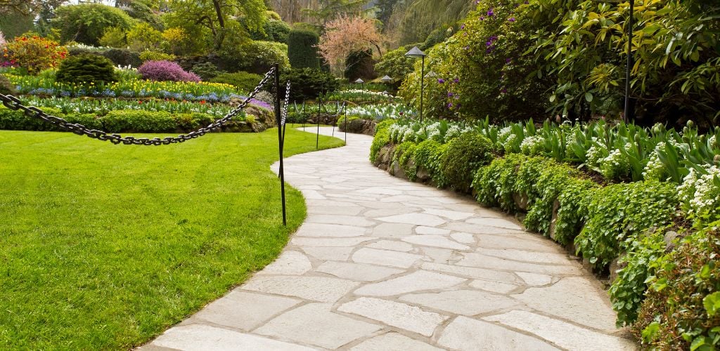 Stone pathway into the garden surrounded by green plants and flowers during daytime. 