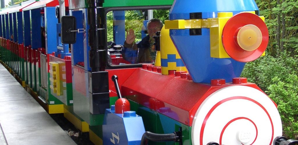 A colorful train looks like a Lego, operated by an old man waving at the camera. 