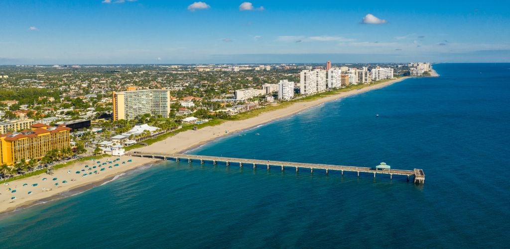 Deerfield Beach, Florida shoreline aerial photo. An island with structures and green plants. Blue tents and blue ocean on the beach. 