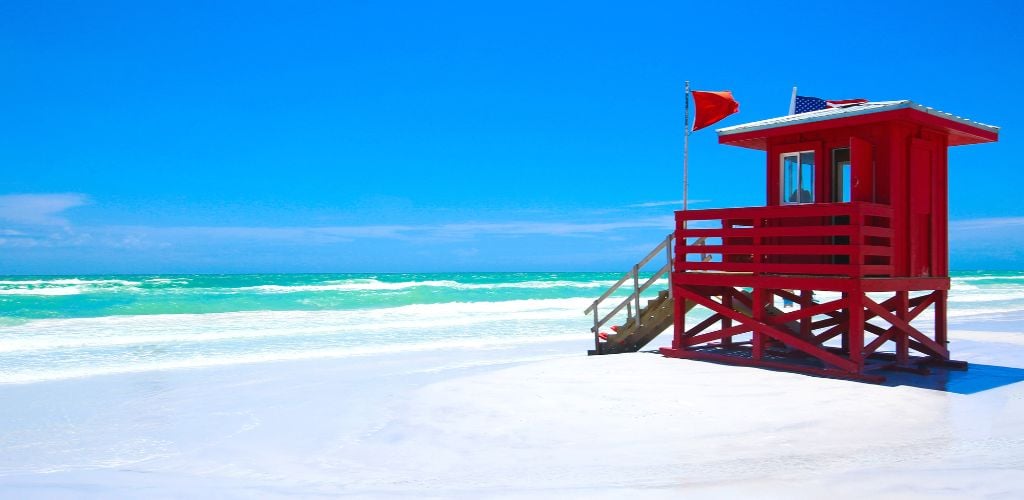 A red lifeguard hut on a lovely beach with white sand, blue sky, and clear water. 