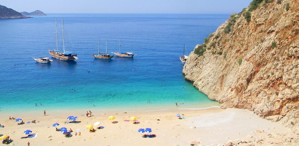 A tiny tent and tourists enjoying the sun on the beach, as well as a four-boat park near the beach. A gorgeous, clear sea with a rocky peak on the horizon. 