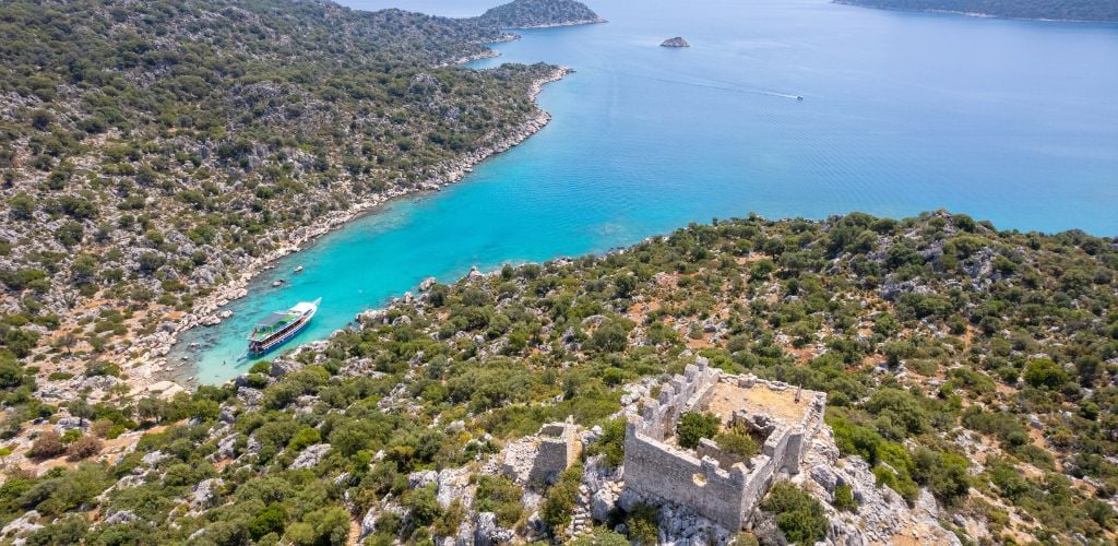 Aerial view of the island with green trees and a big boat parked near shore and blue sea. An old castle on the top of the hill. 