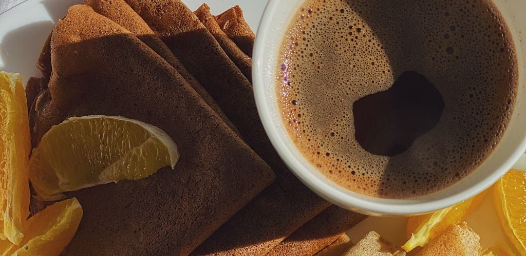 A Cup of Coffee Beside Crepes and orange. 