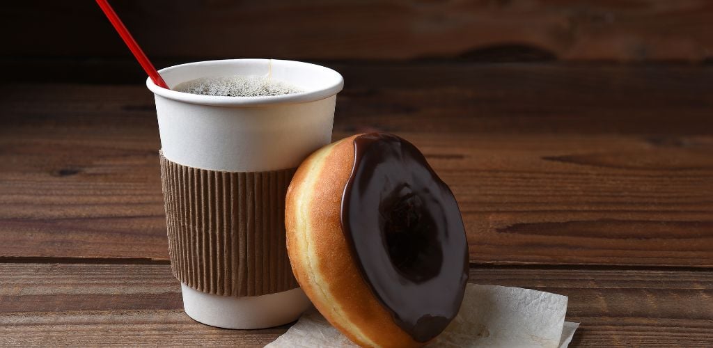 A chocolate frosted donut leaning on a hot cup of freshly brewed coffee. 