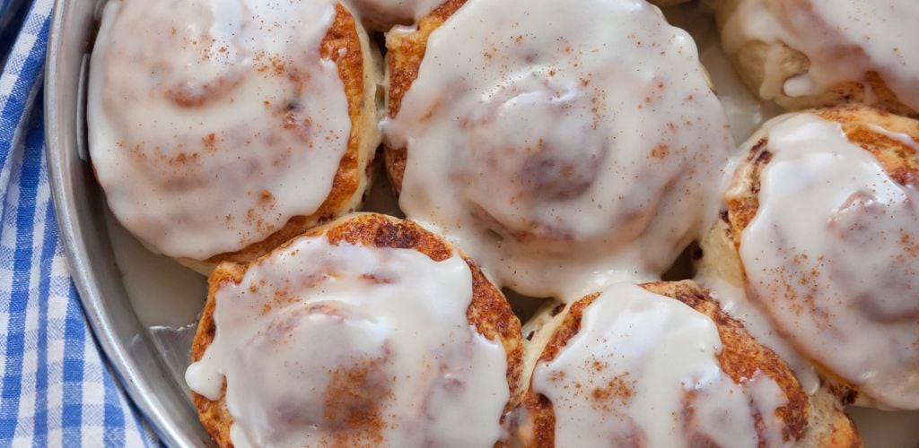 Cinnamon buns sit on a blue and white checked towel, straight from the oven. An icing glaze tops them. 