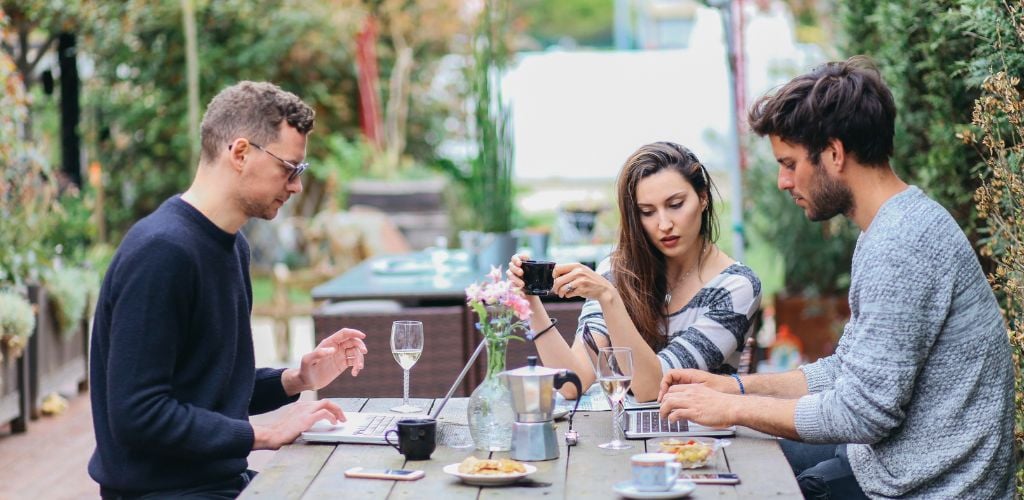 Two men and a woman working outdoor and on the wooden table there is a laptop, a glass of water, and a cup of coffee. 