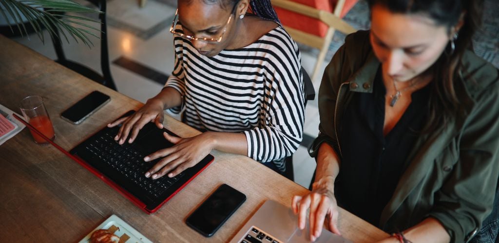 Two young women using a laptop and there is a phone, and a glass of juice.