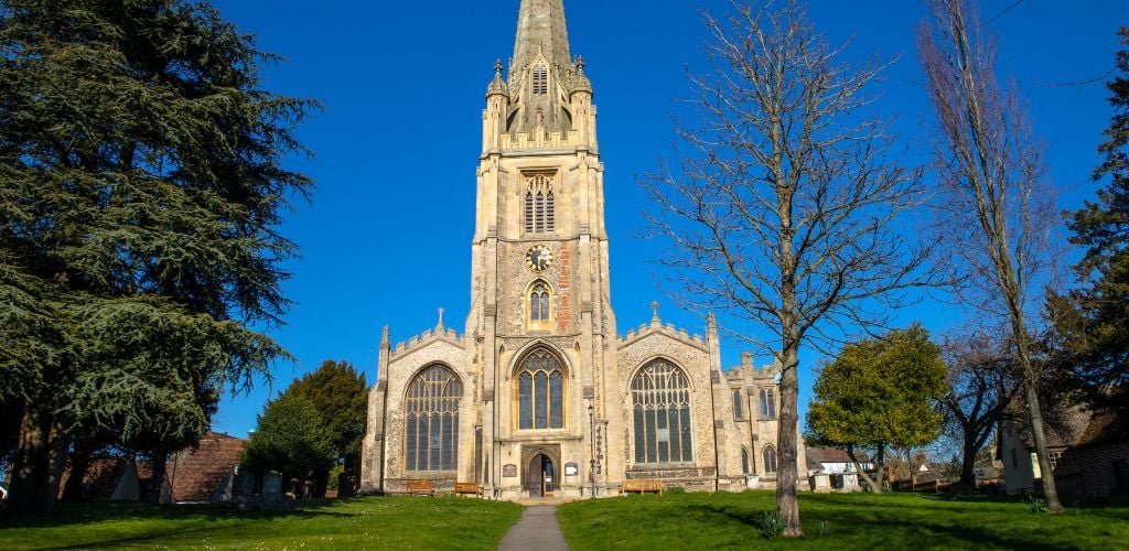 A historic church with a large clock in the center of the structure and green trees and grass on the outside. 